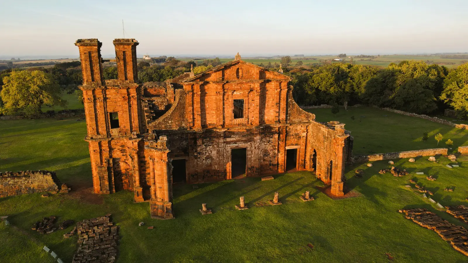 Ruínas da igreja de São Miguel Arcanjo ao entardecer, com fachada de pedra avermelhada em estilo barroco e vasto gramado ao redor.